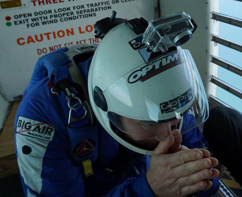 Brian Germain in a helmet, hands pressed together, seated by the open door of an aircraft